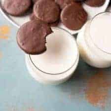 round chocolate cookies on white plate with one cookie sitting on side of top of glass with white milk in it.