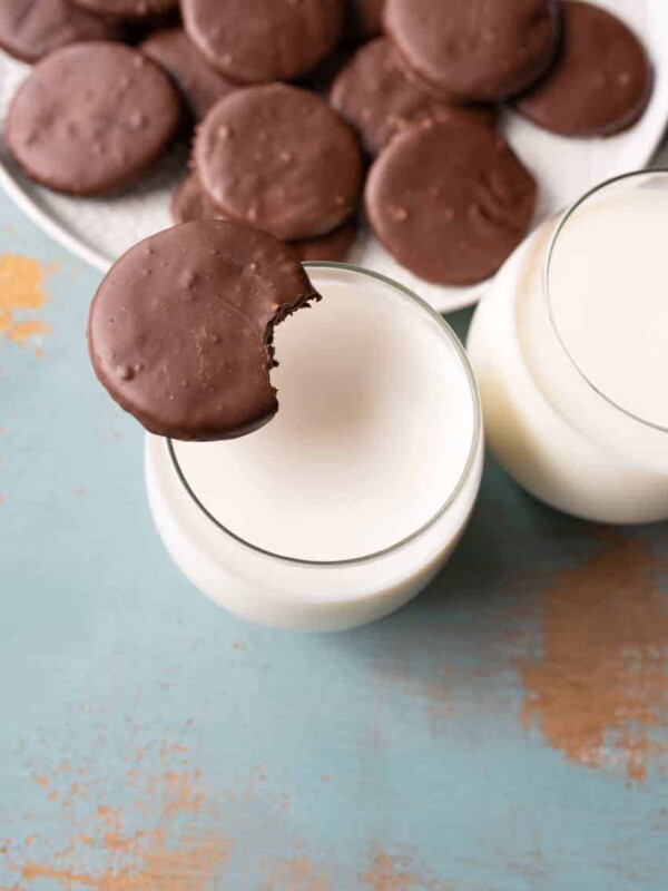 round chocolate cookies on white plate with one cookie sitting on side of top of glass with white milk in it.