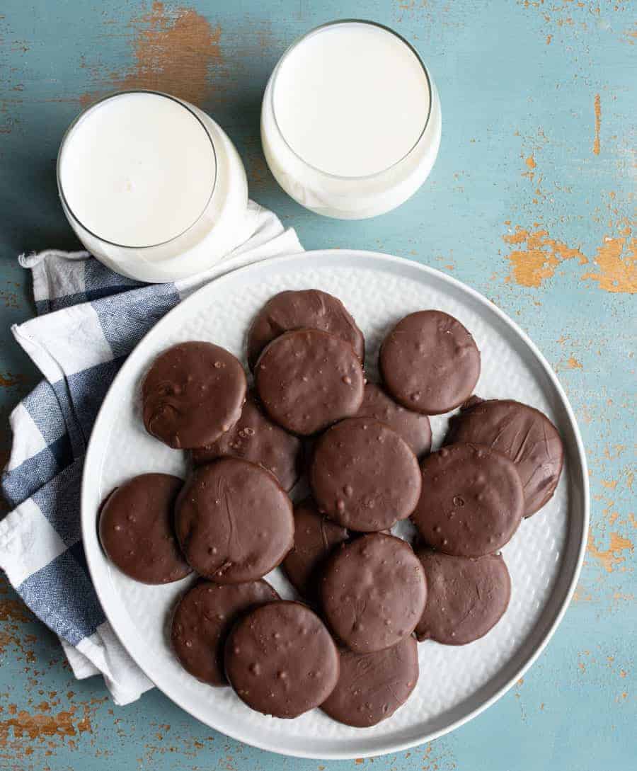 round chocolate cookies on white plate with two glasses of white milk and light blue background.