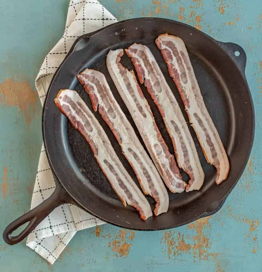 black cast iron skillet on greenish background with black and white towel under edge and uncooked bacon in pan.