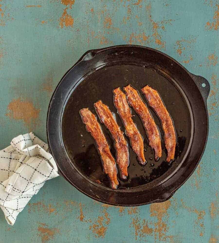 black cast iron skillet on greenish background with a black and white towel around handle and cooked bacon with grease in skillet.