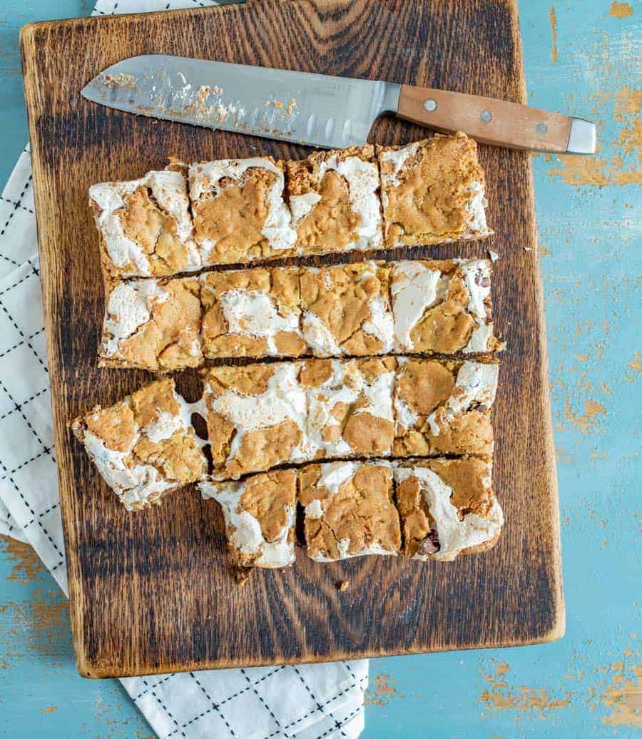 tan and white cookie bars on a brown cutting board on top of a white towel with a brown-handled knife on the board.