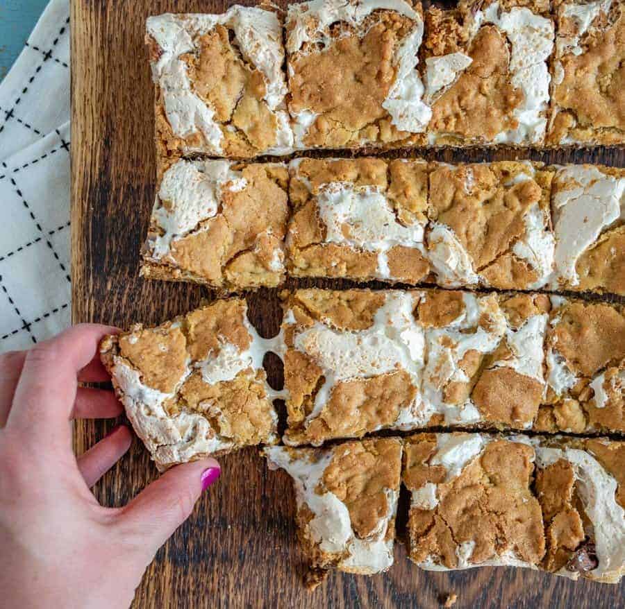 tan and white peanut butter s'mores bars on a brown cutting board on top of a white towel with a hand pulling off one bar.