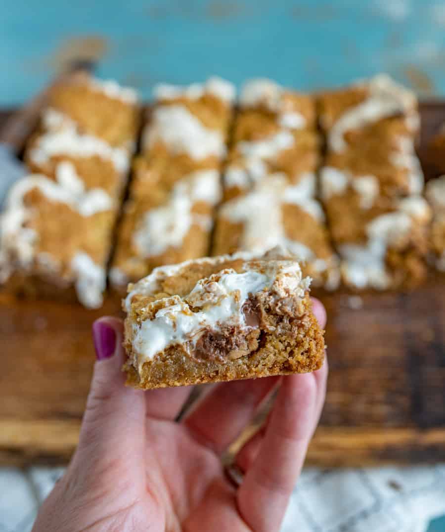 tan and white cookie bars on a brown cutting board on top of a white towel with a hand pulling off one bar.