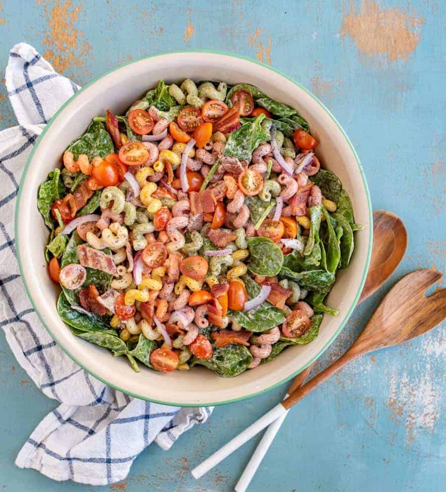 pasta salad with different colored vegetables in a big bowl with wooden spoons sitting on a blue and white towel.