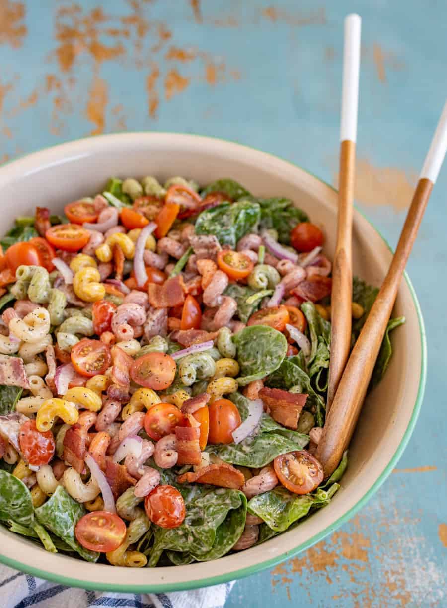 spinach bacon pasta salad in a big bowl with wooden spoons sitting on a blue and white towel.