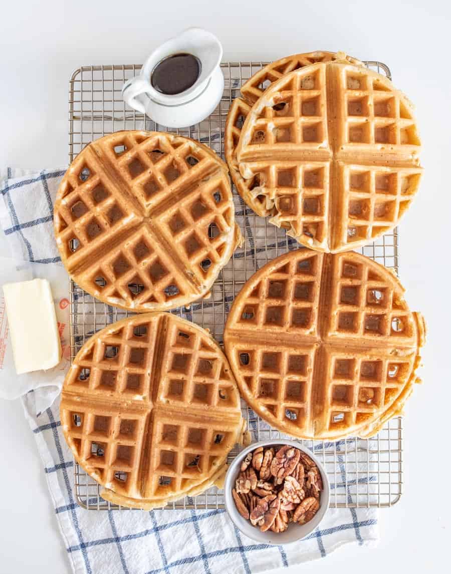 waffles on a wire rack on top of blue and white towel with small bowl of pecans, part of a stick of butter, and white syrup jar.