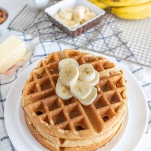 A stack of banana waffles topped with banana slices sits on a white plate. Nearby, theres a block of butter, an extra bowl of banana slices, a bunch of whole bananas, and a milk jug on a plaid tablecloth.