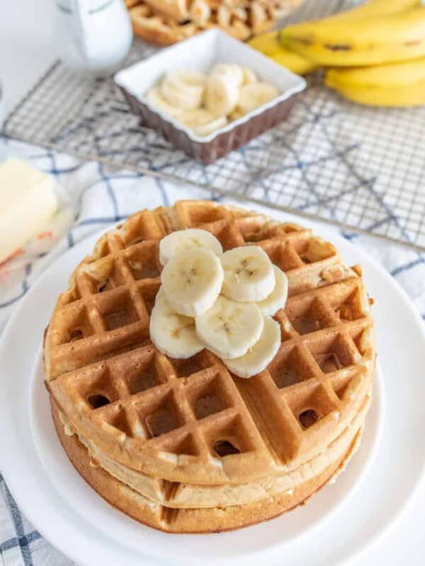 A stack of banana waffles topped with banana slices sits on a white plate. Nearby, theres a block of butter, an extra bowl of banana slices, a bunch of whole bananas, and a milk jug on a plaid tablecloth.