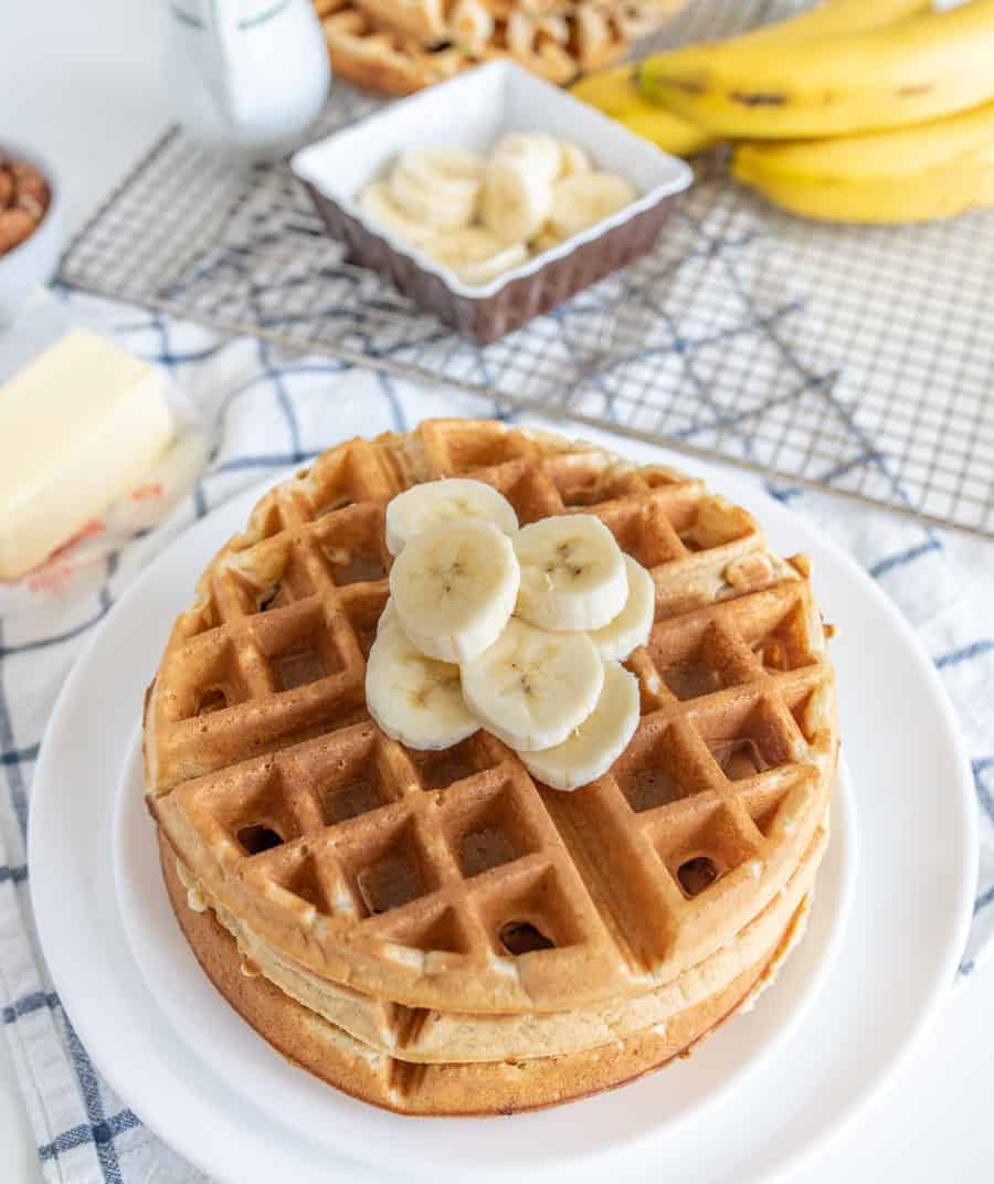 image for Banana Waffles Recipe with waffles stacked on a white plate and sliced bananas on top with sliced bananas, whole bananas, butter, and a white syrup jar in the background.