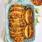 A baking dish holds several cheesy baked chicken tacos filled with meat, beans, and topped with diced tomatoes, onions, and herbs. Lime wedges and a bowl of salsa are on the side. The dish sits on a striped kitchen towel.