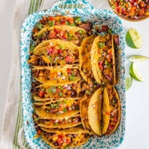 A baking dish holds several cheesy baked chicken tacos filled with meat, beans, and topped with diced tomatoes, onions, and herbs. Lime wedges and a bowl of salsa are on the side. The dish sits on a striped kitchen towel.