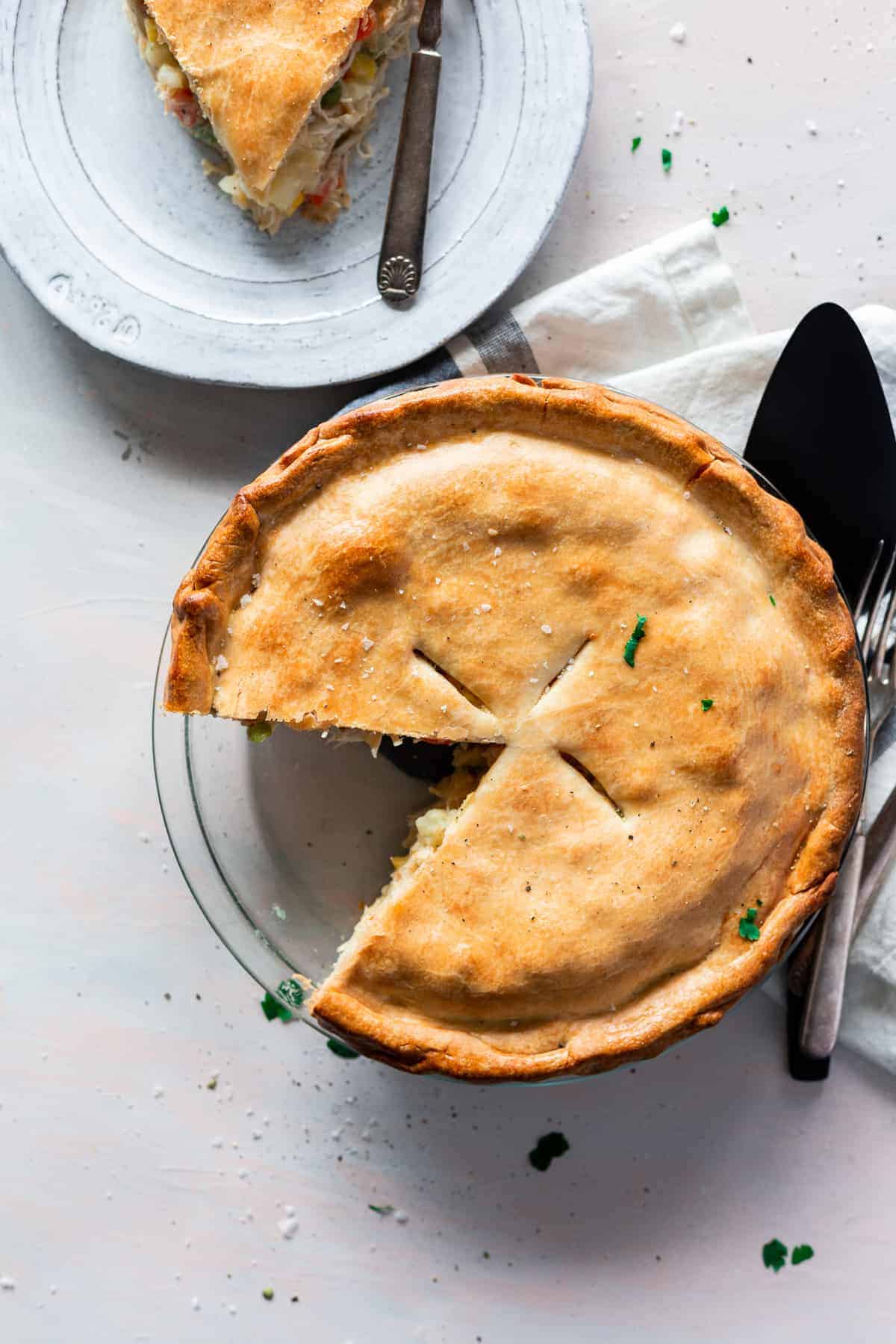 Chicken pot pie in a glass pie dish with one piece cut out and on a white plate behind it.