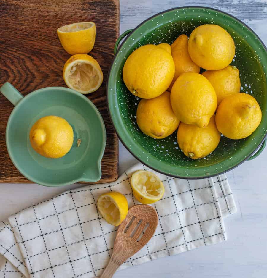 A cutting board with cut lemon halves and a lemon juicer next to a green colander filled with fresh yellow lemons