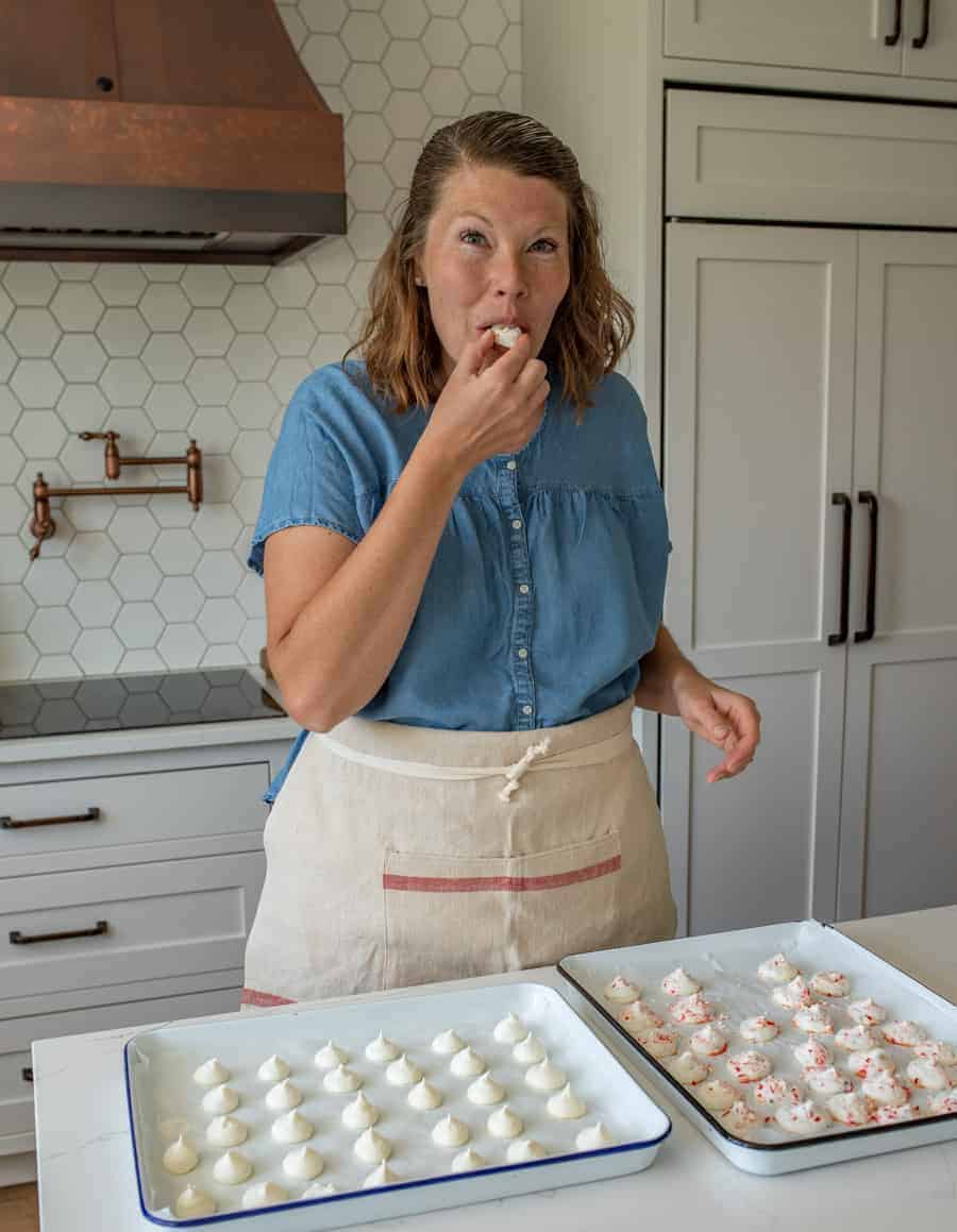 Lady in blue shirt and apron tasting something two pans of meringue cookies in front of her.