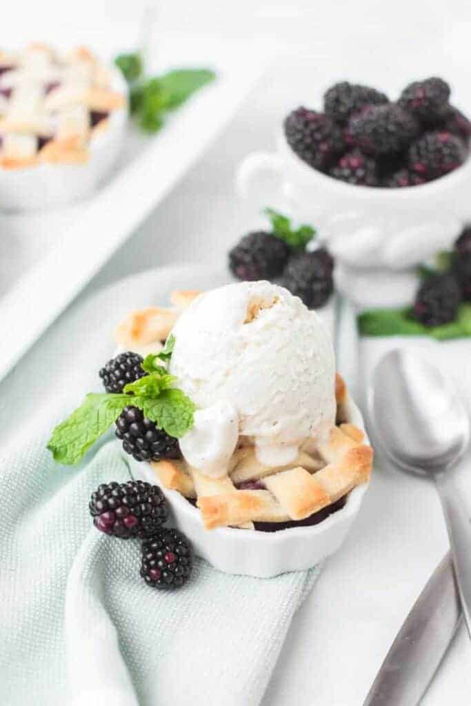 small white dishes filled with blackberry pie filling and a lattice crust with blackberries all around and vanilla ice cream