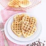 Heart-shaped chocolate chip waffles stacked on a pristine white plate. A red-striped cloth and fork rest to the left, while additional chocolatey delights cool on a wire rack in the background, surrounded by scattered chips for an extra touch of sweetness.
