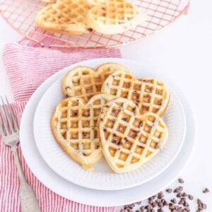 Heart-shaped chocolate chip waffles stacked on a pristine white plate. A red-striped cloth and fork rest to the left, while additional chocolatey delights cool on a wire rack in the background, surrounded by scattered chips for an extra touch of sweetness.