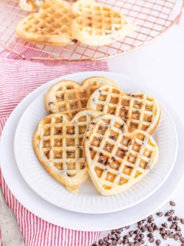 Heart-shaped chocolate chip waffles stacked on a pristine white plate. A red-striped cloth and fork rest to the left, while additional chocolatey delights cool on a wire rack in the background, surrounded by scattered chips for an extra touch of sweetness.