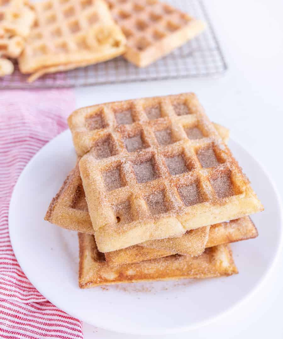 A stack of four waffles dusted with cinnamon sugar on a white plate. A red and white striped cloth is partially visible beside the plate. More waffles are on a cooling rack in the background.
