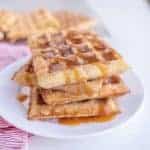 A stack of churro waffles drizzled with caramel sauce sits on a white plate. The waffles are sprinkled with cinnamon sugar, and a striped red and white napkin is visible beside the plate.