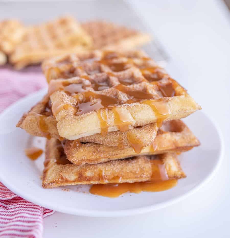 A stack of churro waffles drizzled with caramel sauce sits on a white plate. The waffles are sprinkled with cinnamon sugar, and a striped red and white napkin is visible beside the plate.