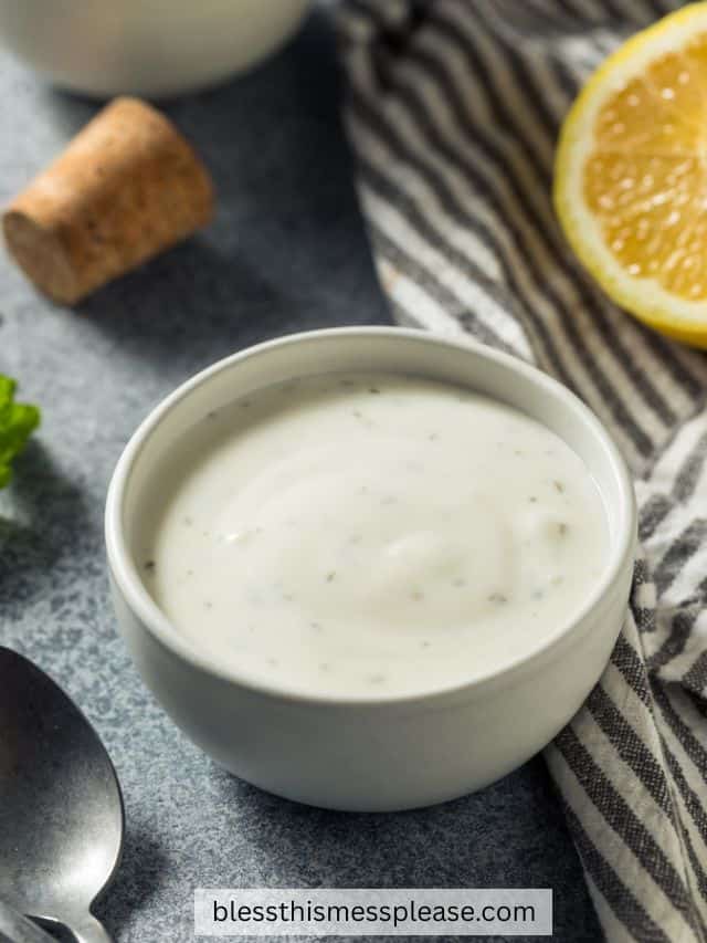A small white bowl filled with creamy ranch dressing sits on a gray surface near a spoon, a cork, a striped cloth, and a lemon wedge—perfect for drizzling over your favorite cornbread salad recipe.