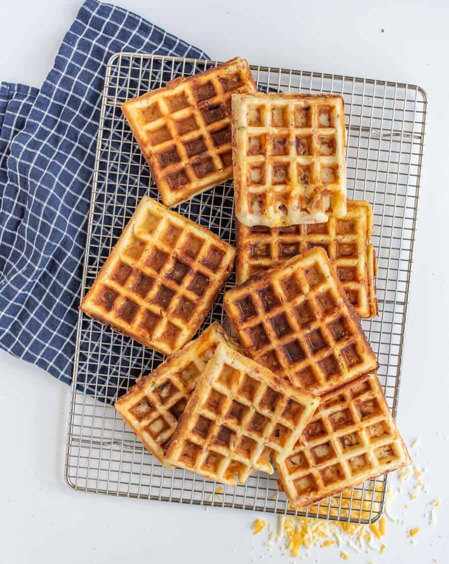 Golden, crisp square waffles stacked randomly on a cooling rack, with a crumpled blue and white checkered cloth beside them. A few crumbs are scattered on the rack, inviting the thought of creamy ice cream melting over their warm surfaces.