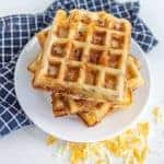 A stack of golden brown, square ham and cheese waffles sits on a white plate, resting on a blue and white checkered cloth. Shredded cheese is scattered around, hinting at the delicious ham and cheese tucked inside these savory waffles.