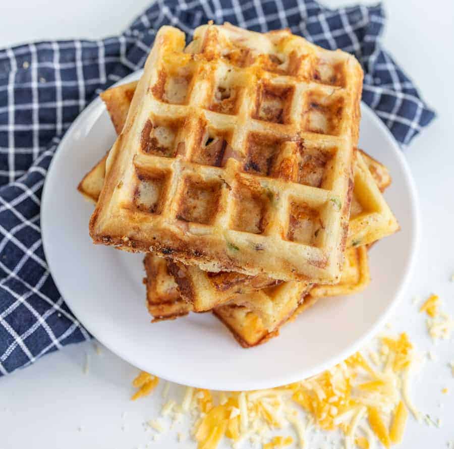 A stack of golden brown, square ham and cheese waffles sits on a white plate, resting on a blue and white checkered cloth. Shredded cheese is scattered around, hinting at the delicious ham and cheese tucked inside these savory waffles.