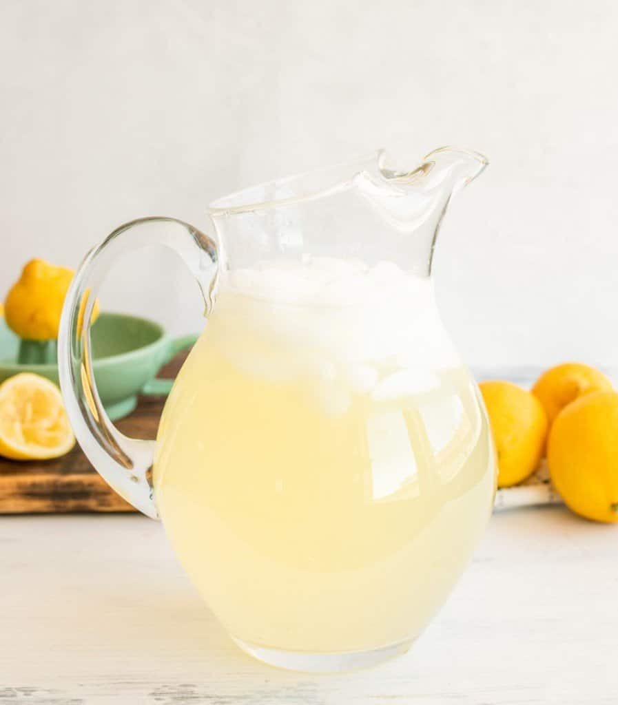 A glass pitcher of light yellow lemonade with ice in it 