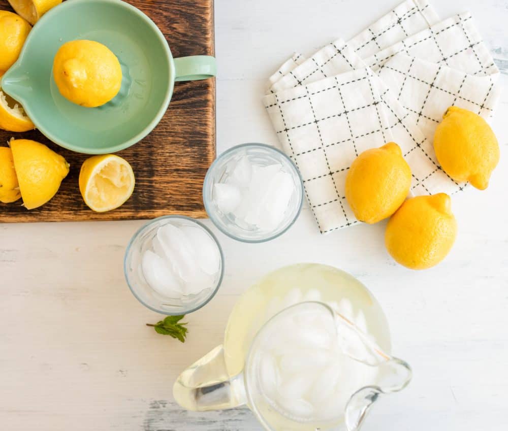 top view of glasses of ice and a pitcher of lemonade with lemons and a ceramic juicer and whole and sliced lemons