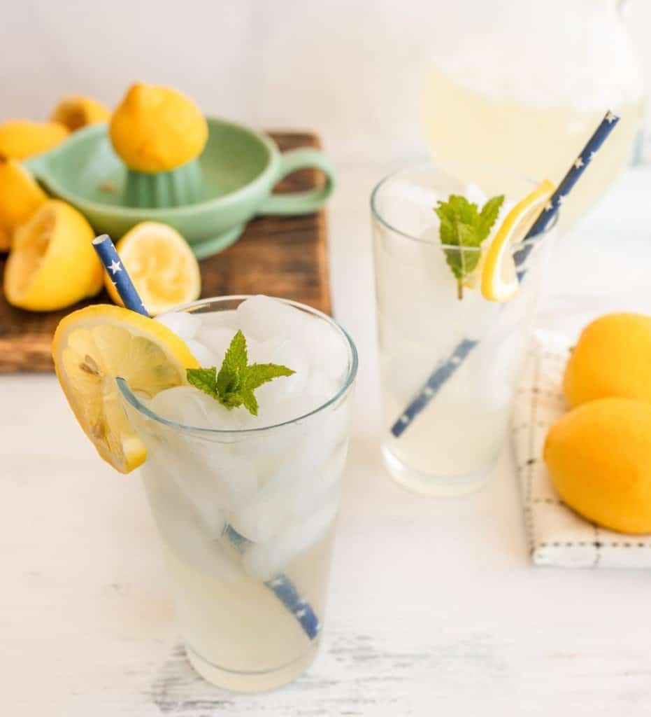 two glasses of lemonade with lemon slices on the rim and straws with the lemon juicer in the back ground