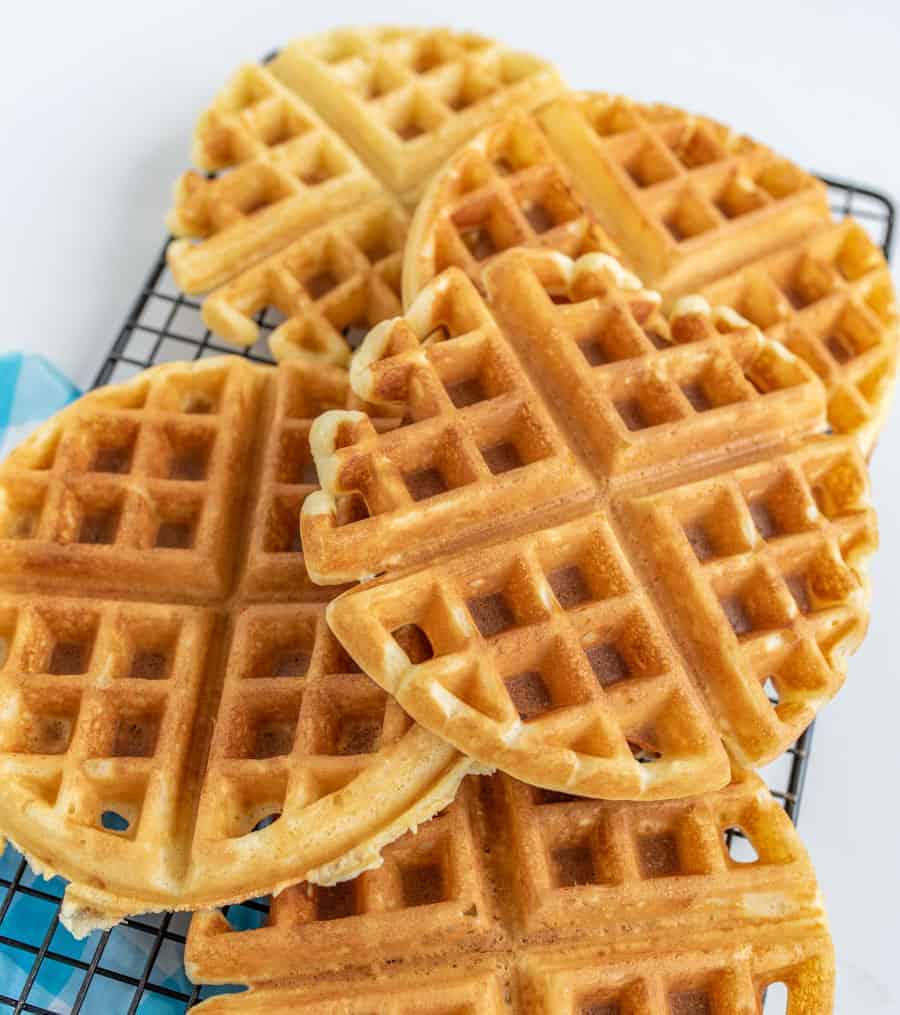 Several waffles stacked and spread out on black wire rack with blue and white towel underneath.