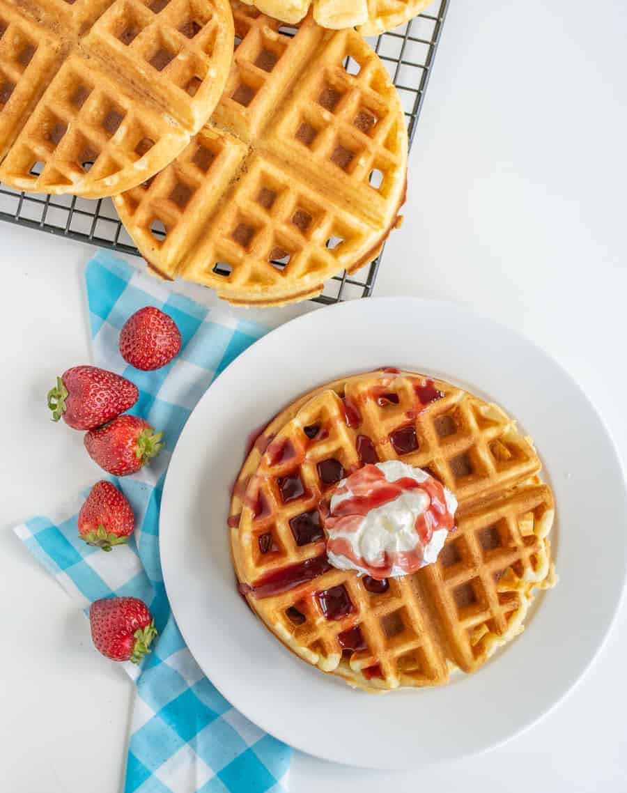 Stack of waffles with cream and red sauce on top and red strawberries next to plate.