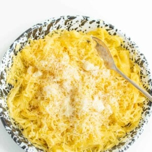 A bowl of cooked spaghetti squash topped with grated cheese and a few dollops of butter, served with a metal fork on a white and black speckled plate.