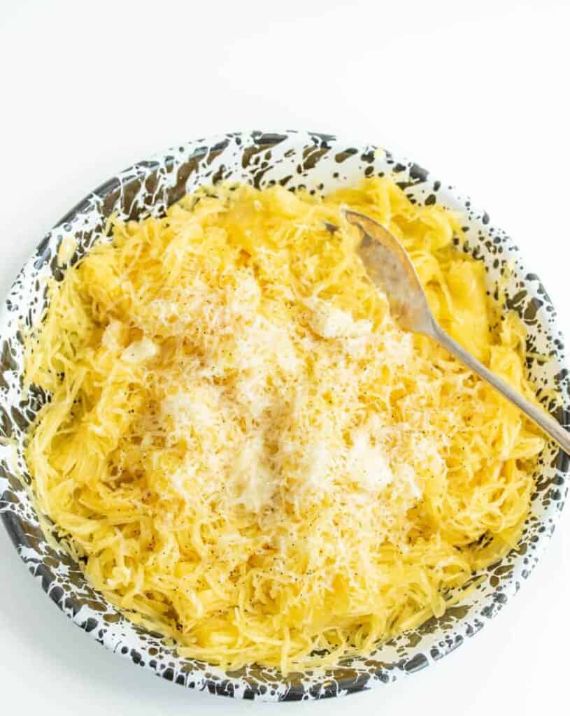 A bowl of cooked spaghetti squash topped with grated cheese and a few dollops of butter, served with a metal fork on a white and black speckled plate.