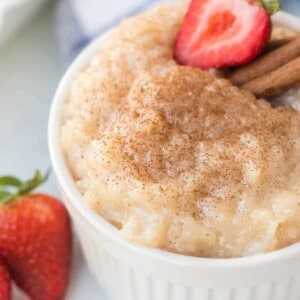 A white bowl filled with the best rice pudding recipe, topped with cinnamon, a sliced strawberry, and cinnamon sticks, with whole strawberries beside the bowl.