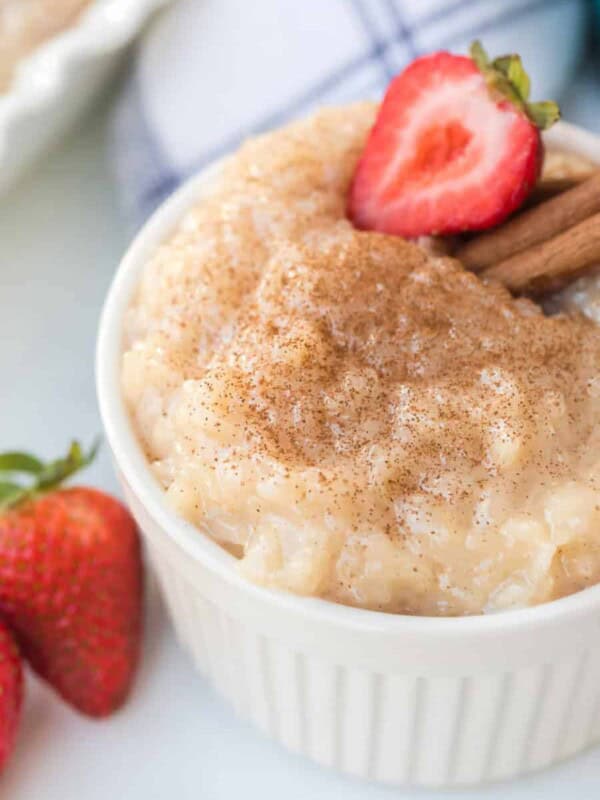 A white bowl filled with the best rice pudding recipe, topped with cinnamon, a sliced strawberry, and cinnamon sticks, with whole strawberries beside the bowl.