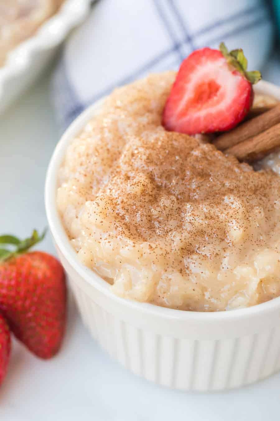 Rice pudding in white ramekin with strawberries and cinnamon sticks.