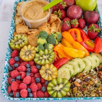 after school snack tray on a blue speckled sheet with mixed fruits, veggies, nuts, hummus and pita chips
