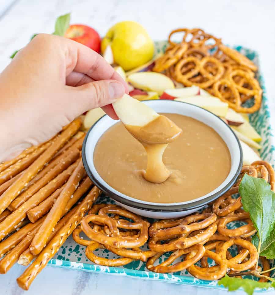 pov hand dipping an apple in the bowl of caramel dip with apple slices and pretzels all around