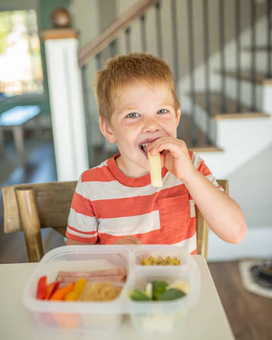 young boy with red hair in red and white striped tee shirt eating a cheese stick with tupperware container with assorted foods in front of him on a table.