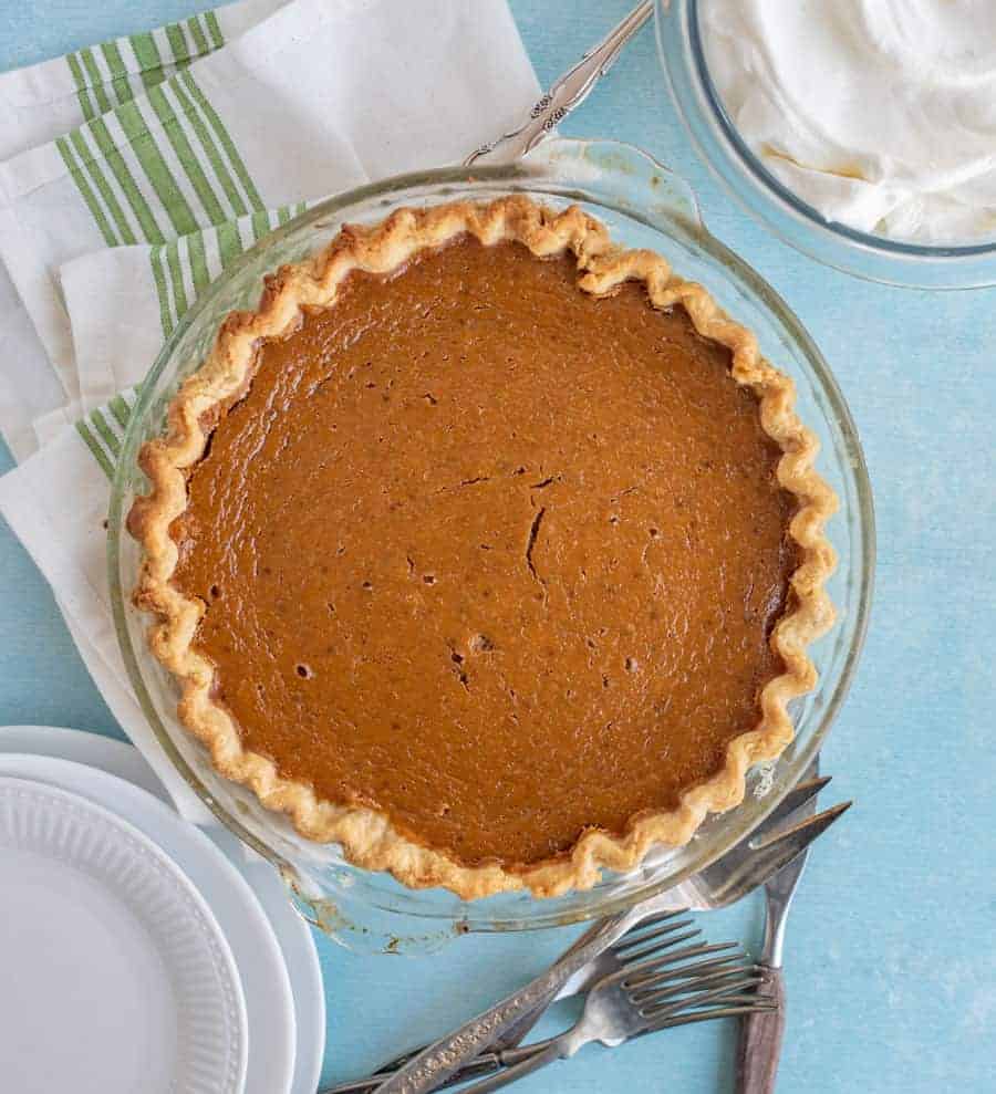 Overhead shot of a baked pumpkin pie in a glass pie dish on a blue counter. A glass bowl with whipping cream is in the top right corner. In the bottom right are some white dessert plates and at the bottom of the pie are some silver forks.