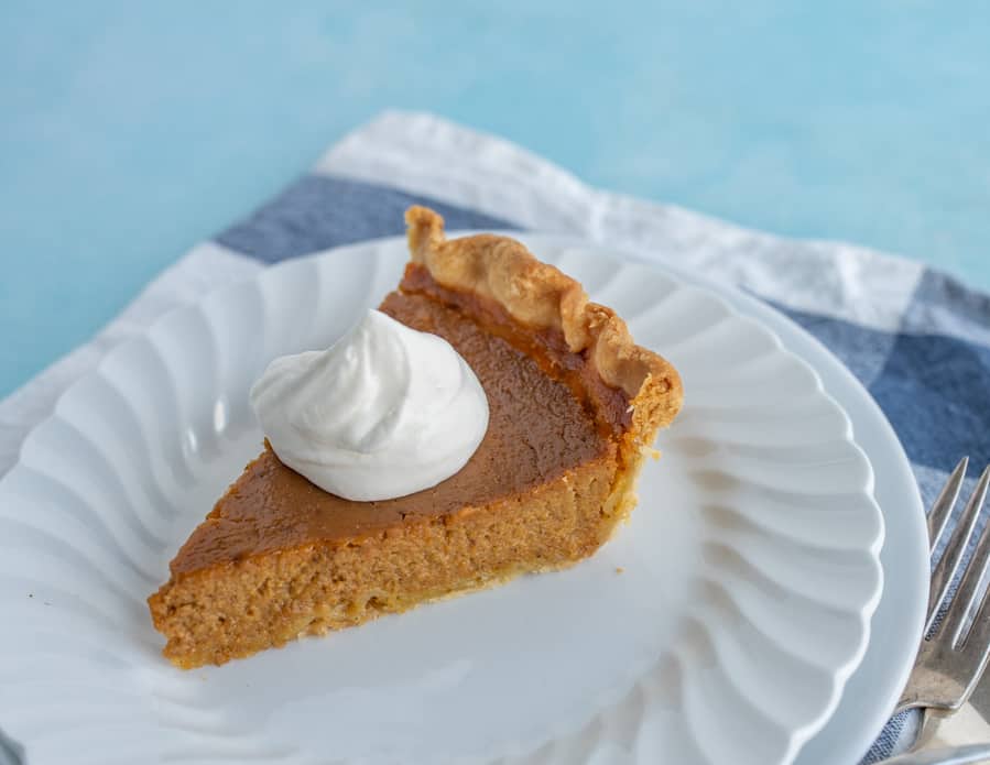 A piece of pumpkin pie from the side on a white dessert plate with a dollop of whipping cream on top.