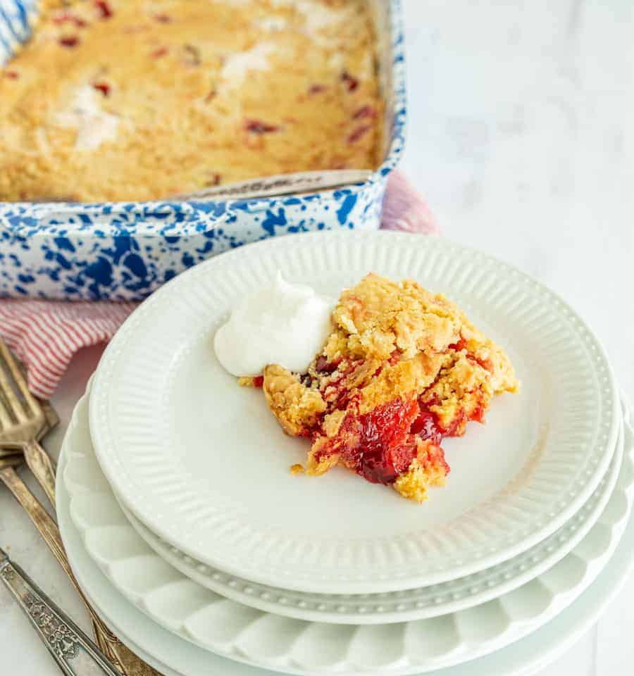 In the foreground is a slice of cherry dump cake with some whipping cream and in the background in the upper left is the pan holding the rest of the cake.