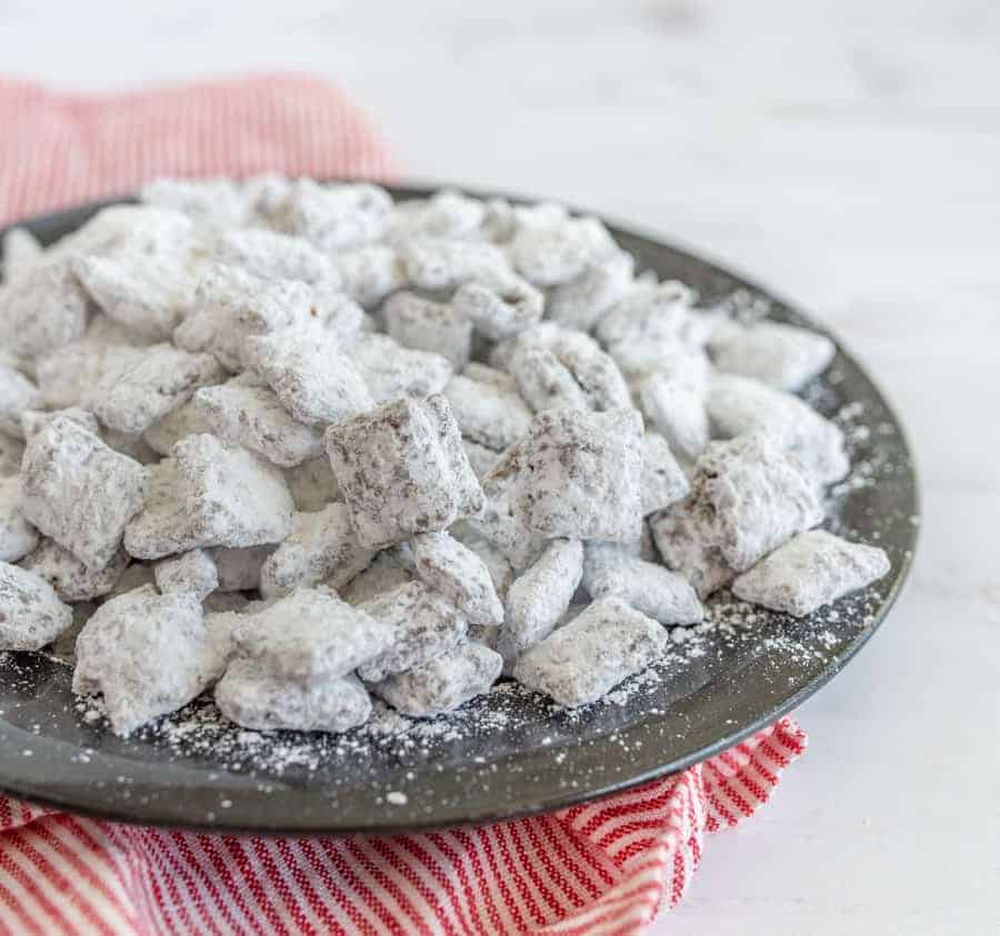 a mixture of chex cereal covered in peanut butter and chocolate and coated with white powdered sugar on a metal plate red and white striped towel in background.