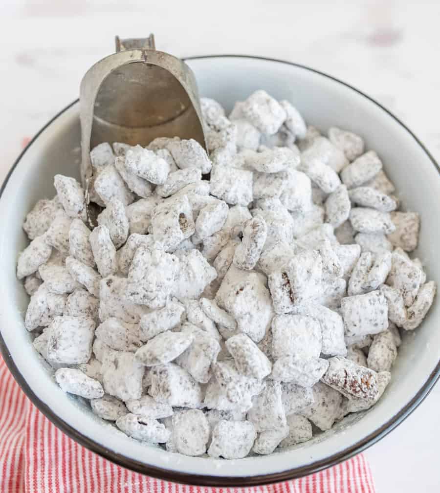 Puppy Chow Recipe a mixture of chex cereal covered in peanut butter and chocolate and coated with white powdered sugar in a white metal bowl with black rim Silver metal scoop in bowl red and white towel in background.