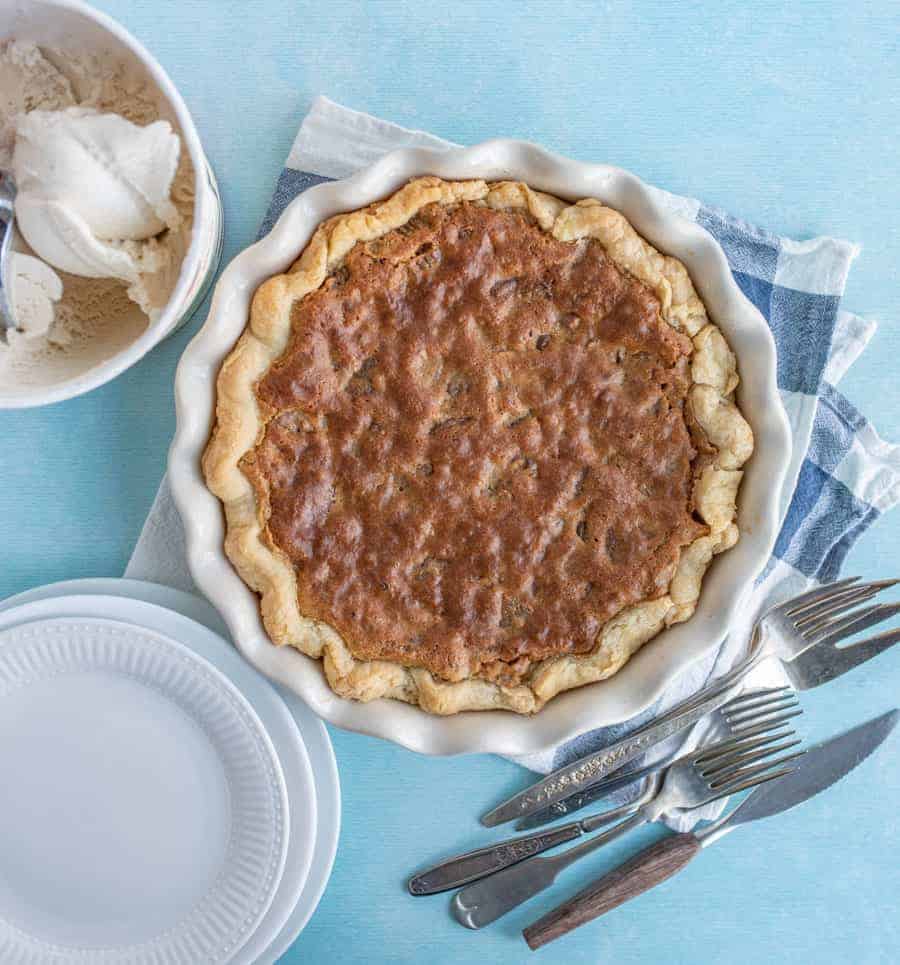 In the middle of the image is a pie inside of a white pie dish that is wavy. The crust matches the waves on the dish and inside the crust is a browned filling. Under the pie dish is a blue and white checkered towel with forks and a pie cutter on it. In the upper left hand corner is a white bowl of ice cream and in the bottom left corner are some empty pie plates.