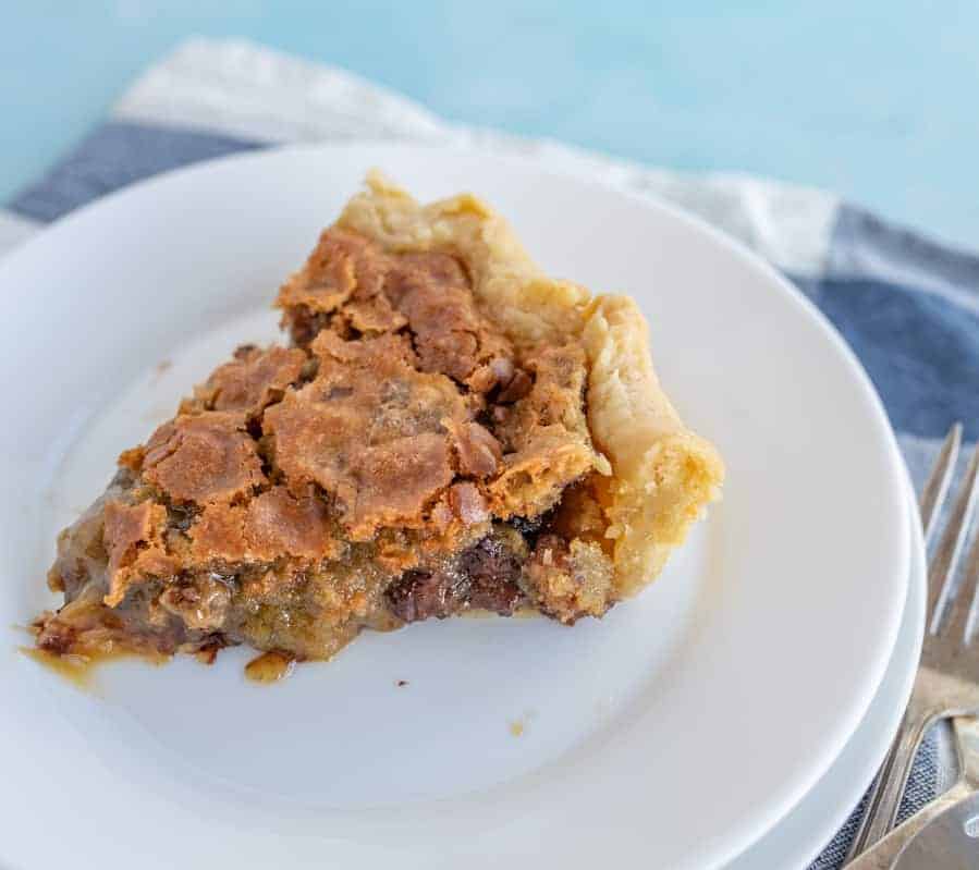 A piece of pie with gooey insides is on a white pie plate with a blue and white checkered towel sticking out from underneath it and a couple of forks on the bottom right hand side are peeking out from under the plate.